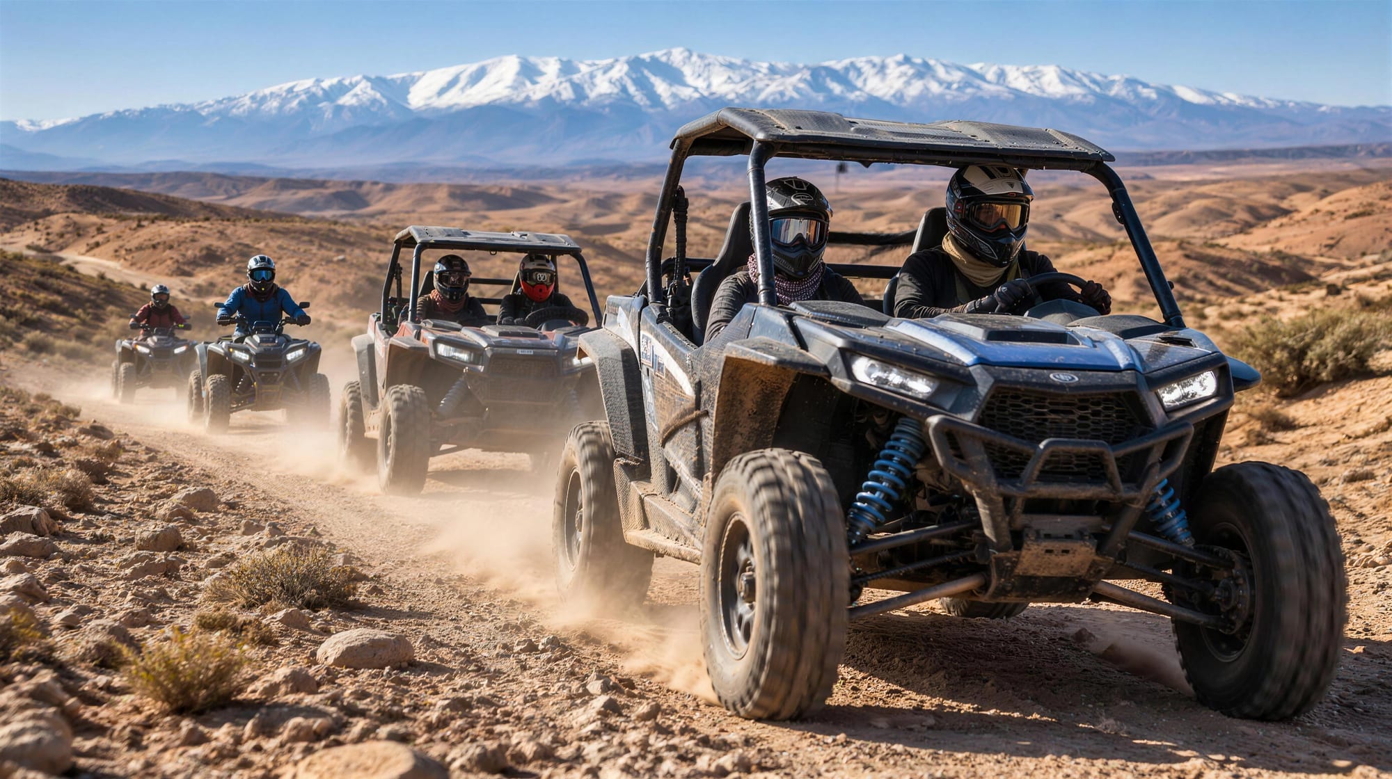 Deux buggys et deux quads en file sur une piste poussiéreuse, pilotes casqués, montagnes enneigées à l’horizon