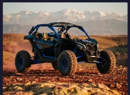 Buggy Can-Am bleu de trois quarts sur un sol rouge, sommets enneigés à l’arrière