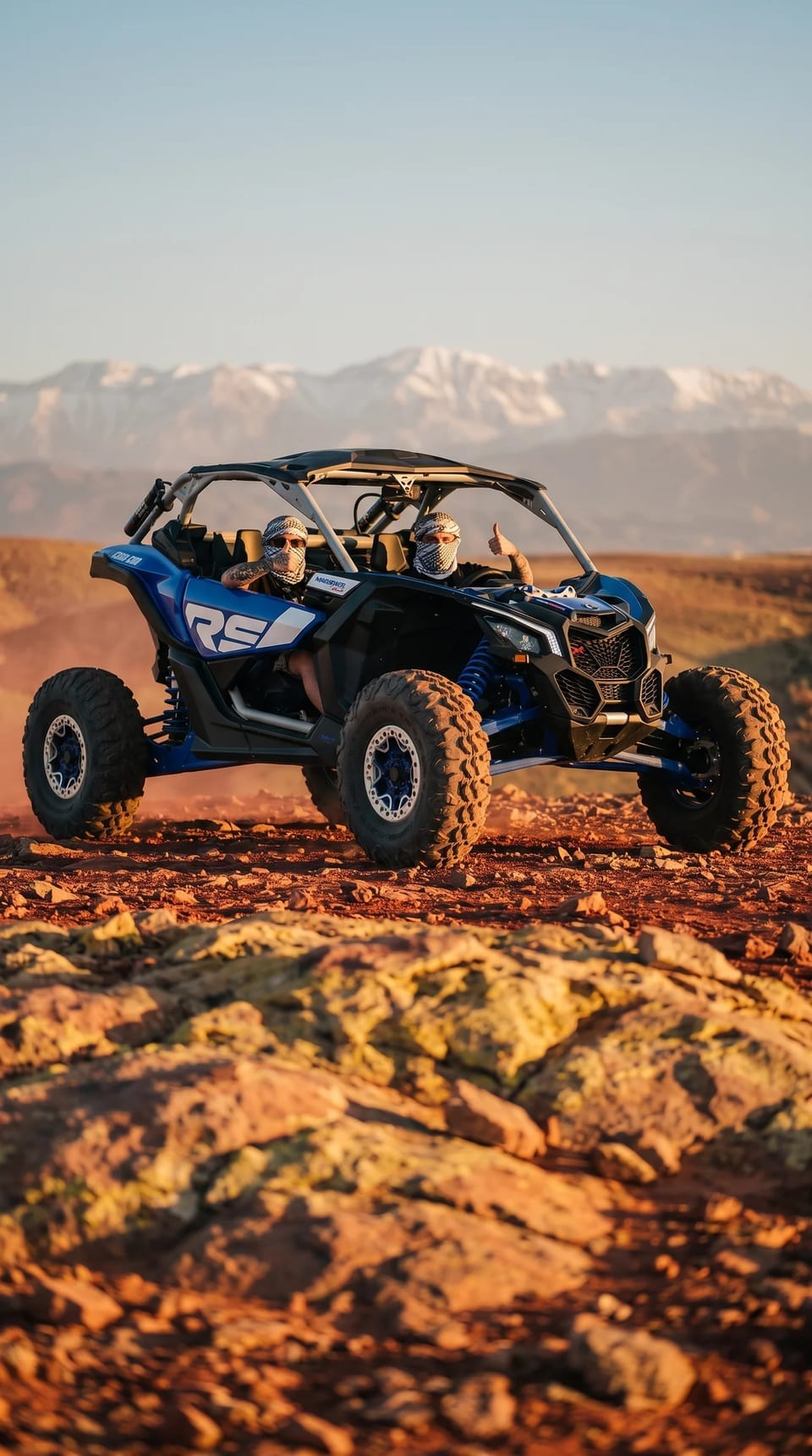 Buggy Can-Am bleu et ses deux pilotes sur un plateau rocheux, Atlas enneigé au fond