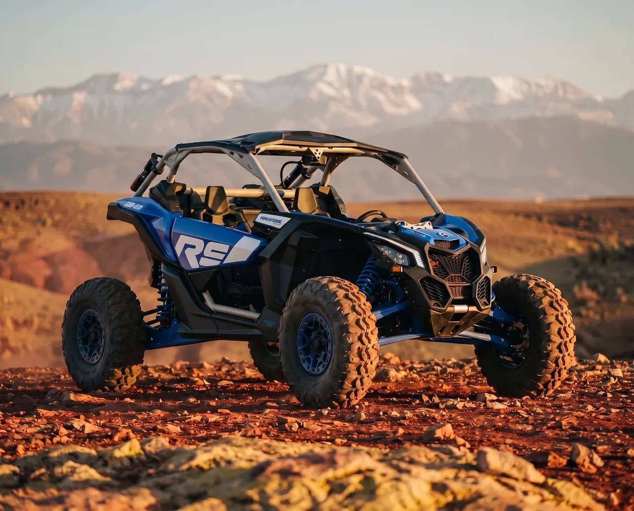 Buggy Can-Am Maverick X3 bleu à l’arrêt sur un sol rocailleux rouge, sommets enneigés au fond