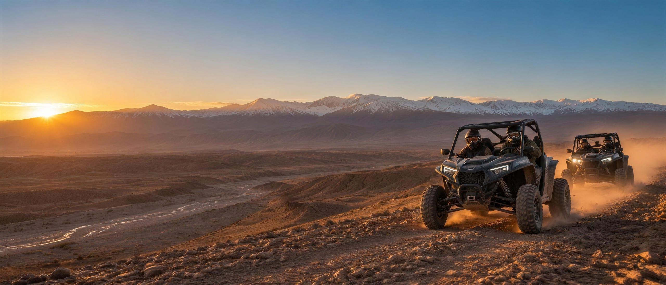Deux buggys soulevant la poussière sur une crête d’Agafay au coucher du soleil, Haut Atlas enneigé à l’horizon